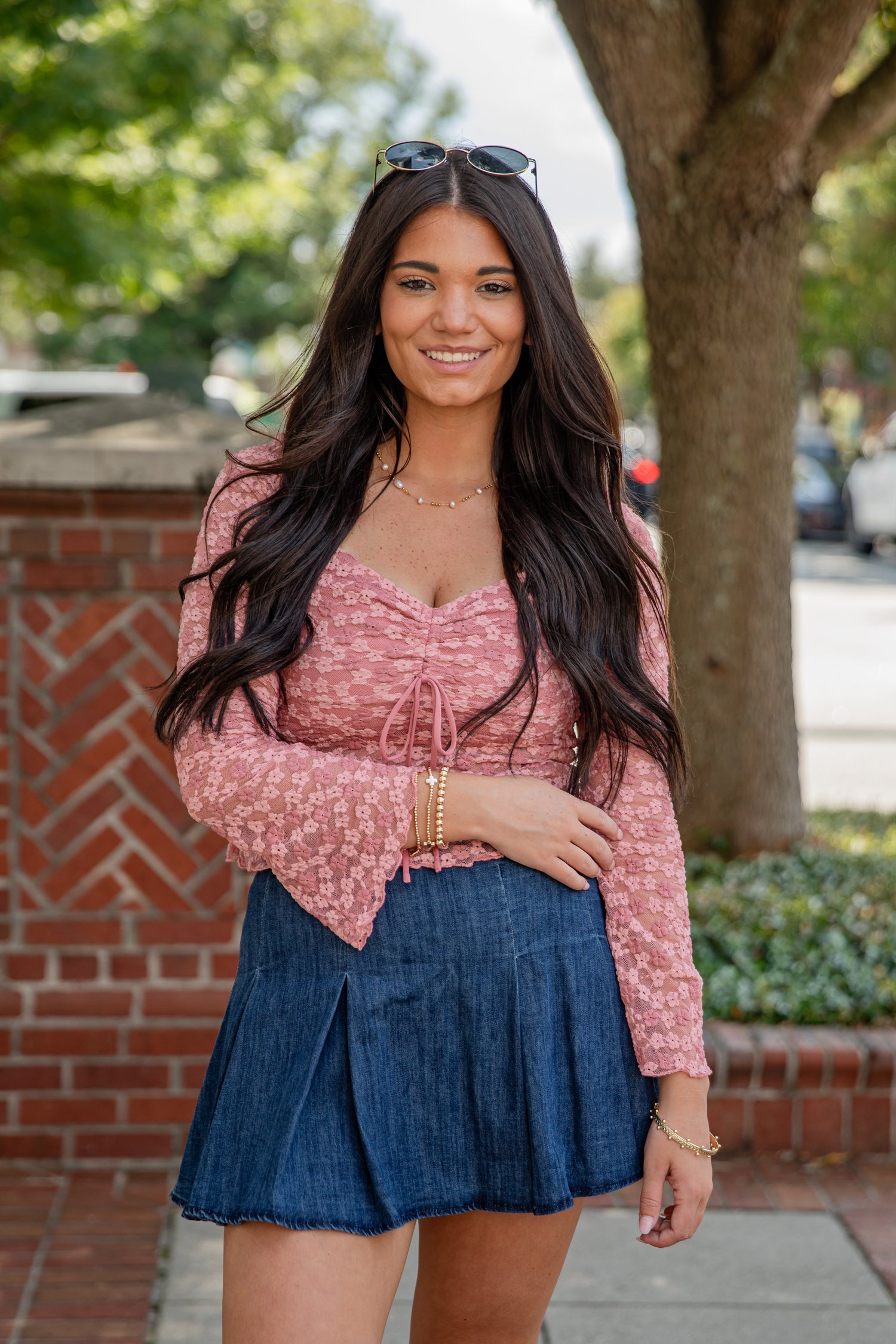 Woman wearing a pink top and blue skirt standing outdoors with a brick wall and tree in the background.