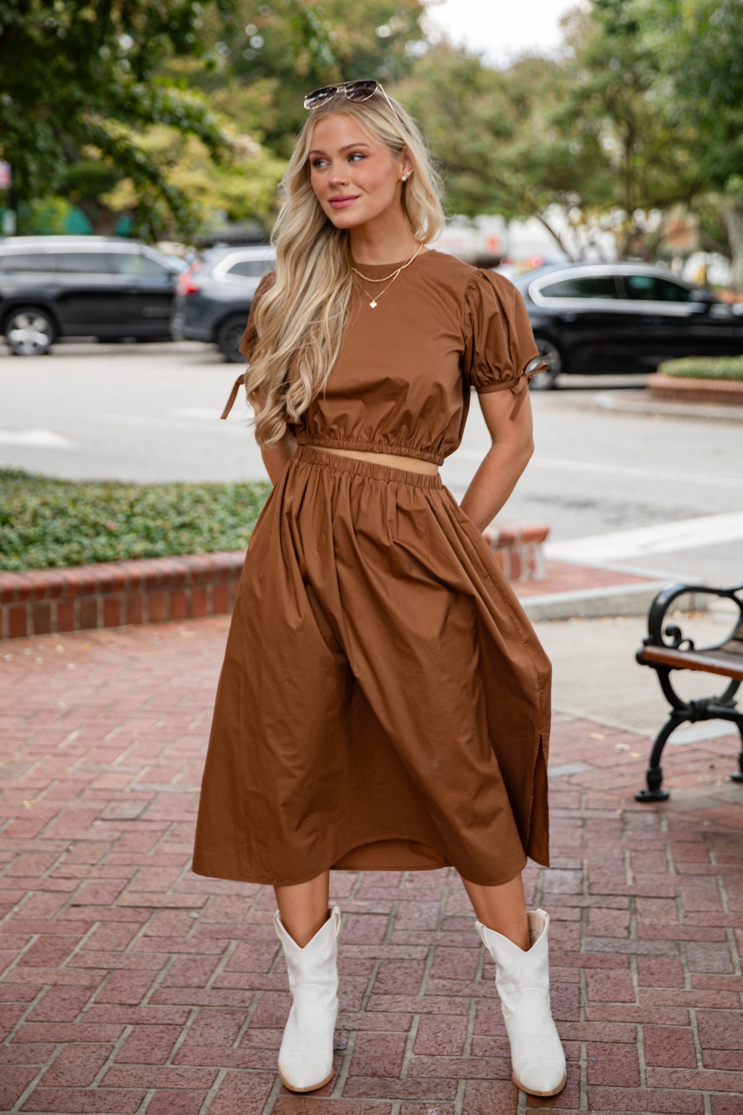 Woman in a brown dress standing on a sidewalk with cars and trees in the background