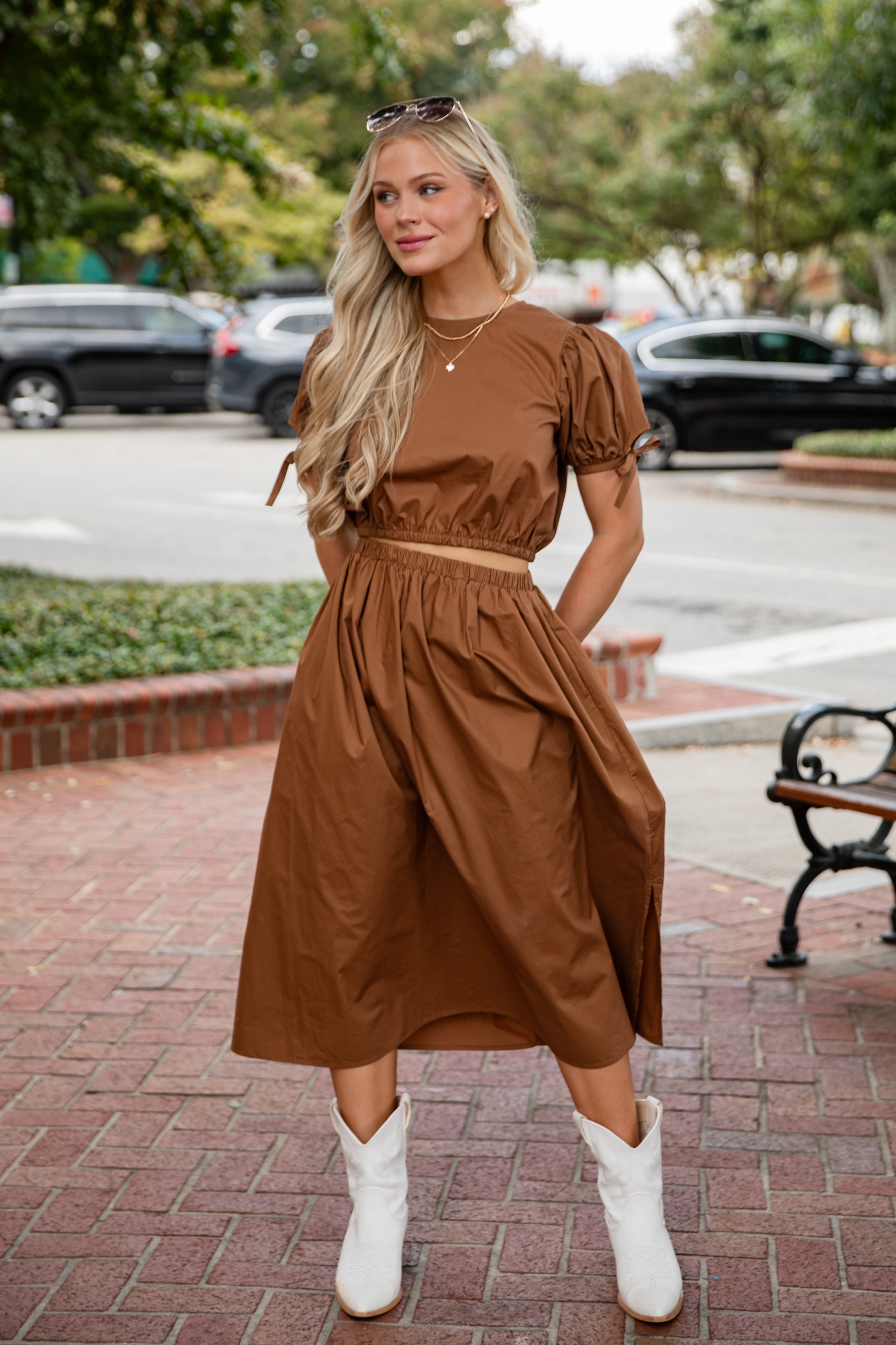 Woman in a brown dress standing on a sidewalk with cars and trees in the background