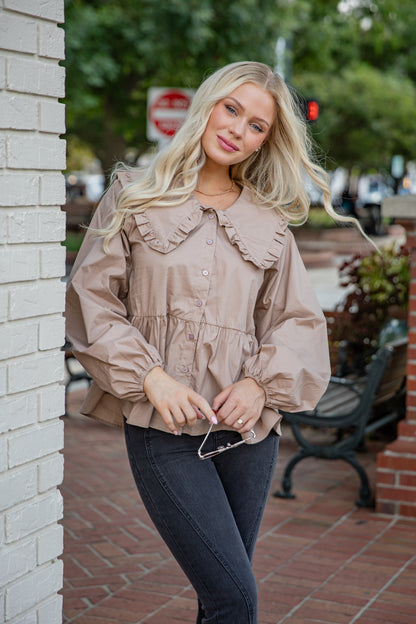 Woman in a beige blouse and dark jeans standing against a brick wall with greenery in the background.