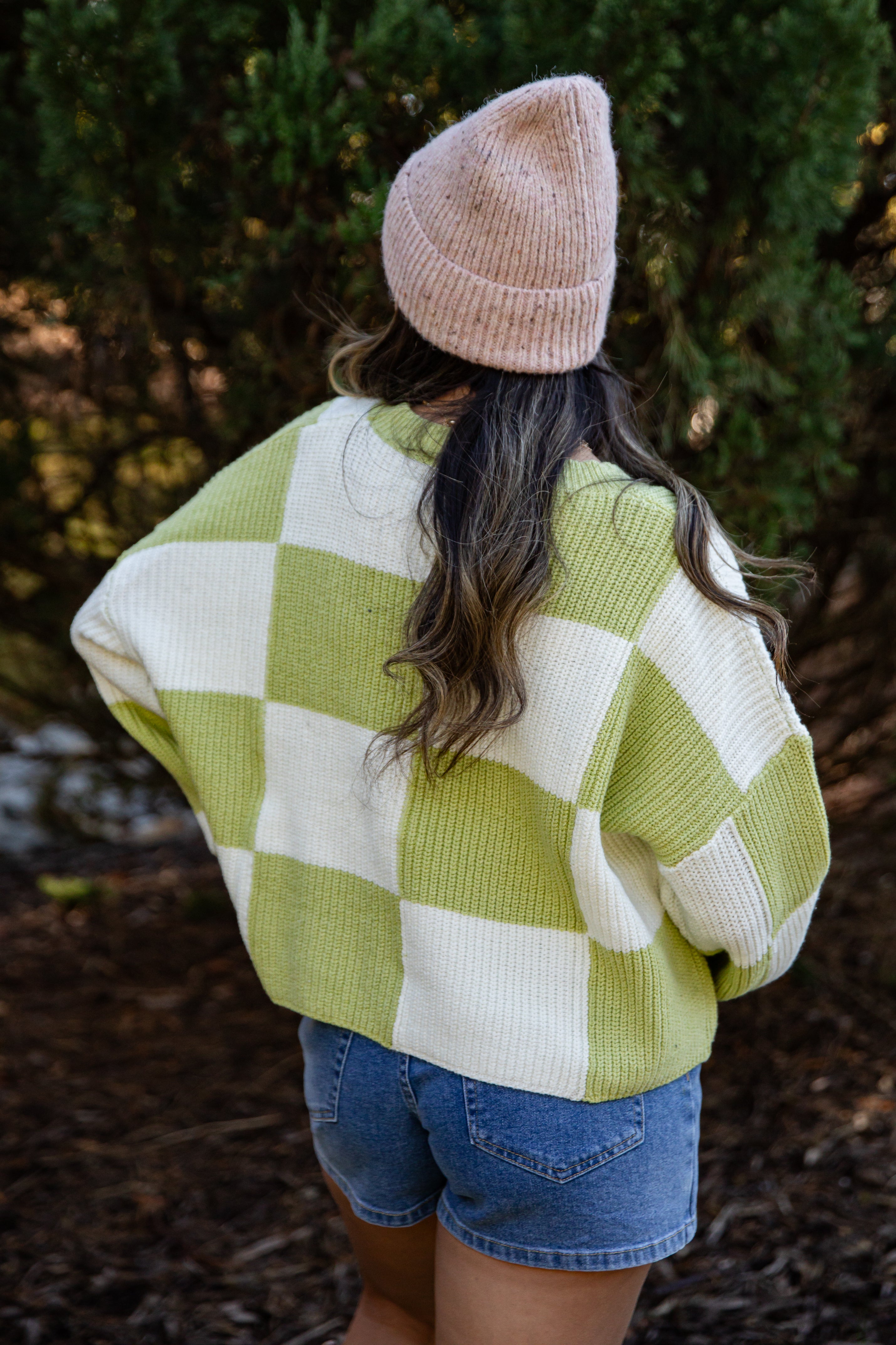 Person wearing a green and white checkered sweater and denim shorts with a beige beanie, standing outdoors.