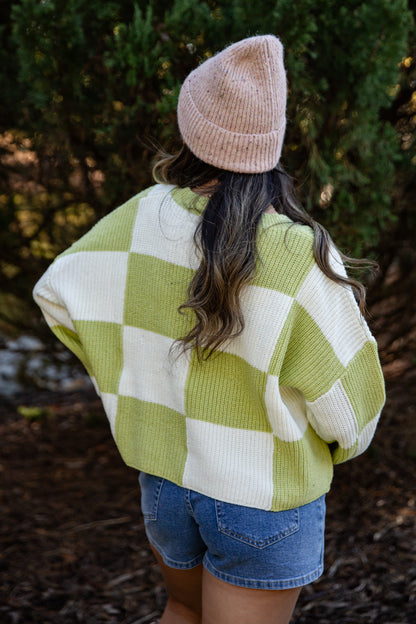 Person wearing a green and white checkered sweater and denim shorts with a beige beanie, standing outdoors.