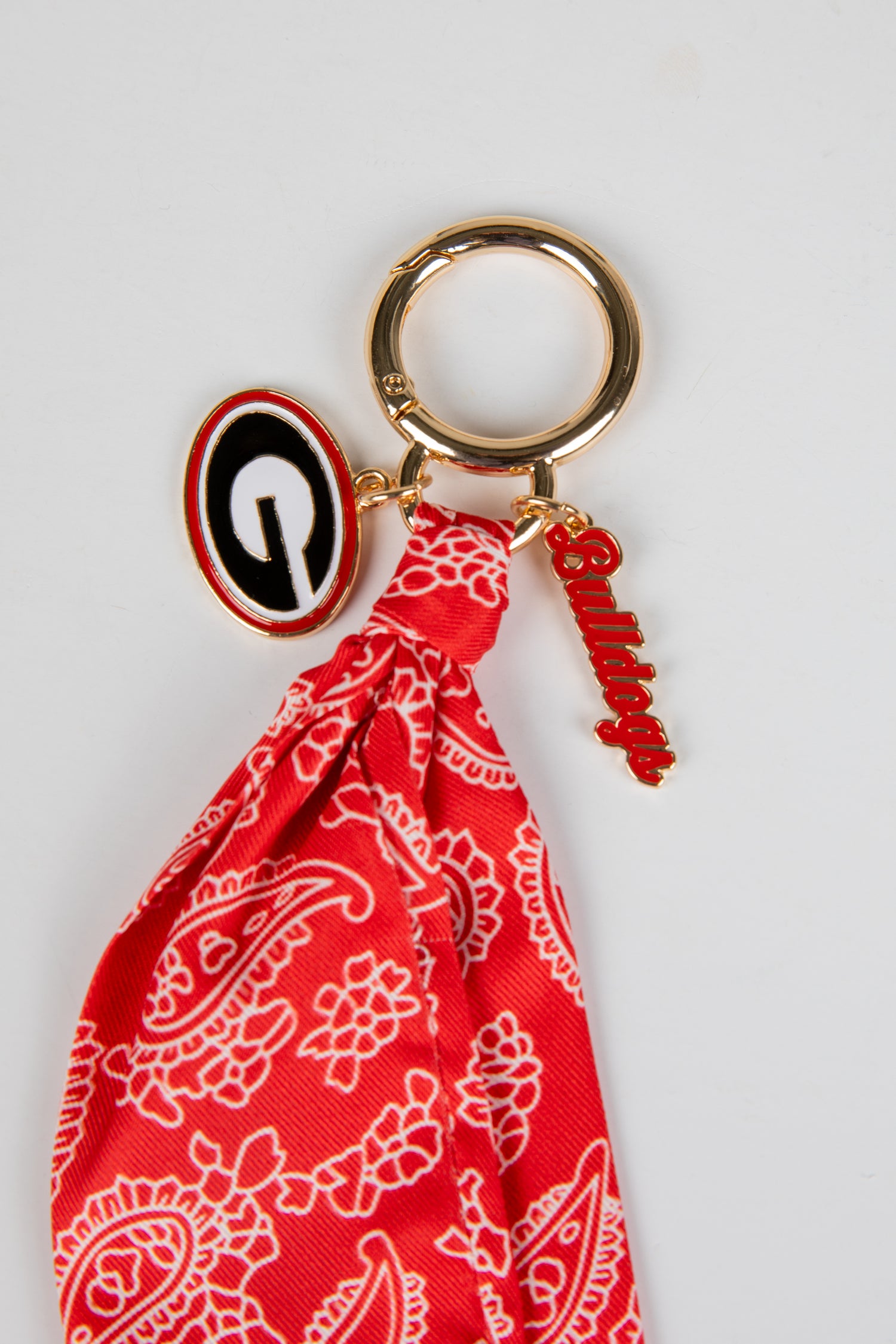 Red bandana keychain with a gold ring and black and white emblem on a light gray background