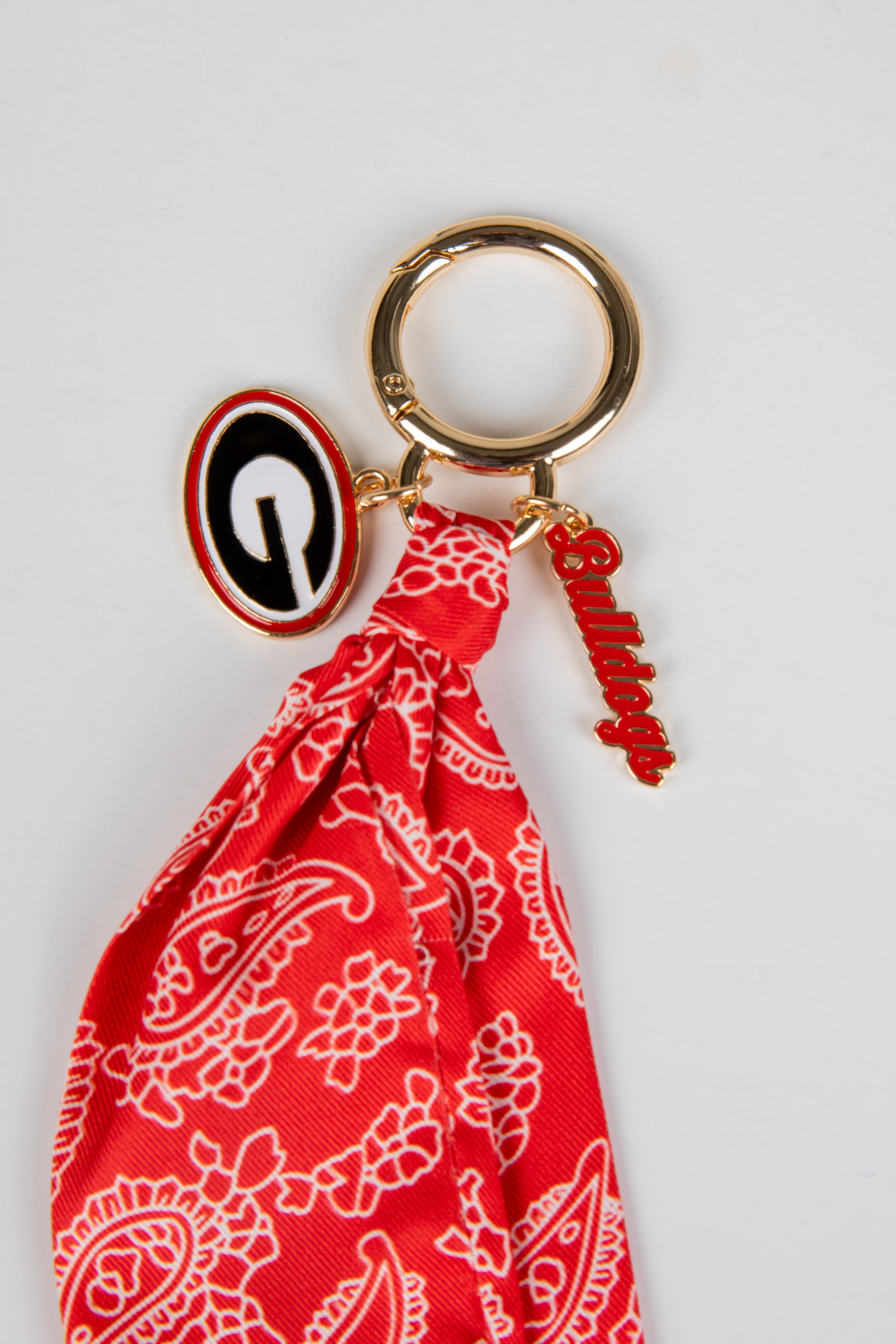 Red bandana keychain with a gold ring and black and white emblem on a light gray background