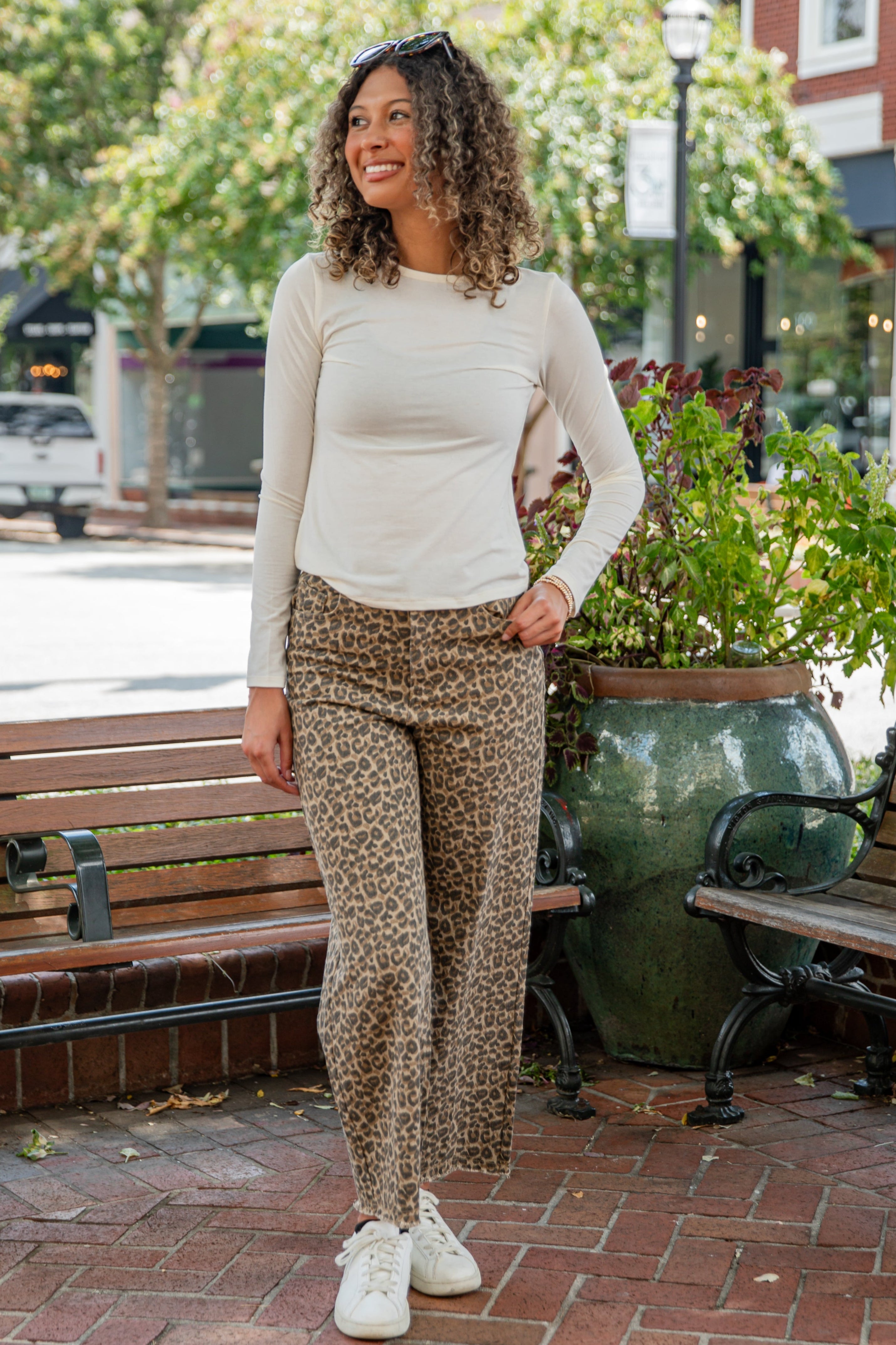 Woman in a white top and patterned pants standing on a sidewalk with plants and benches in the background.