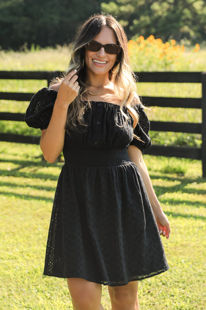 Woman in a black dress standing in a grassy field with a wooden fence and trees in the background.