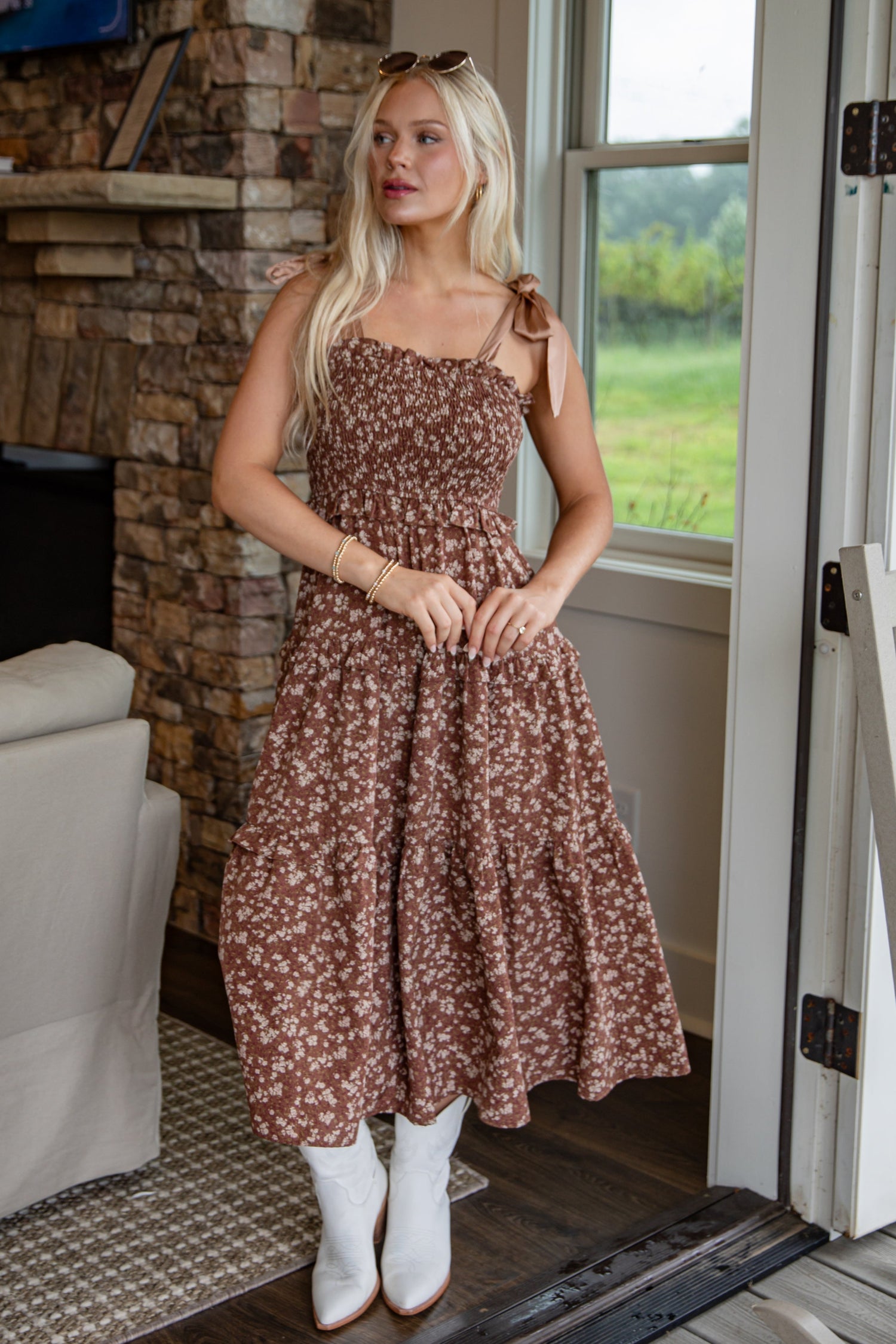 Woman in a floral dress standing in a doorway with a stone fireplace and greenery outside.