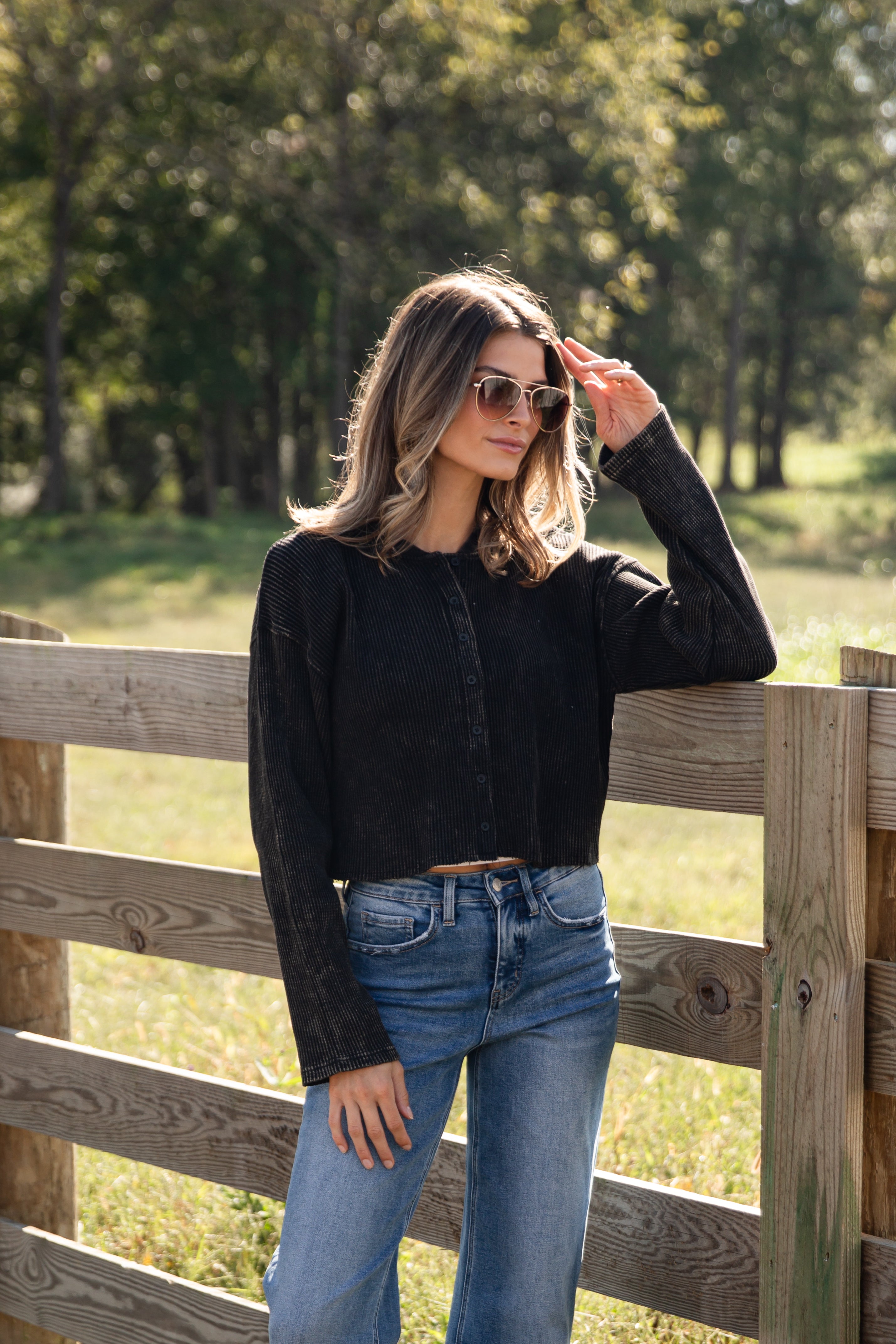 Woman wearing sunglasses and a black jacket standing by a wooden fence with trees in the background