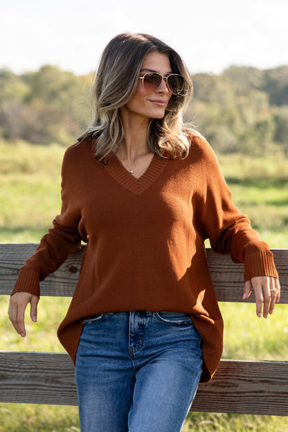Woman in a brown sweater and blue jeans sitting on a wooden fence in a field.