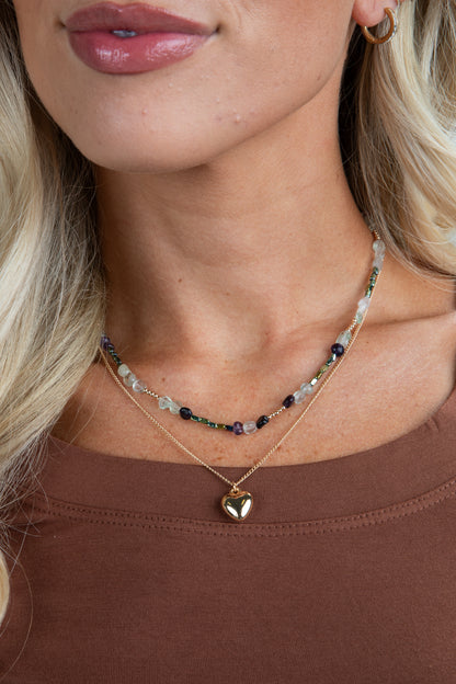 Close-up of a woman wearing a gold heart-shaped pendant necklace with a brown top.