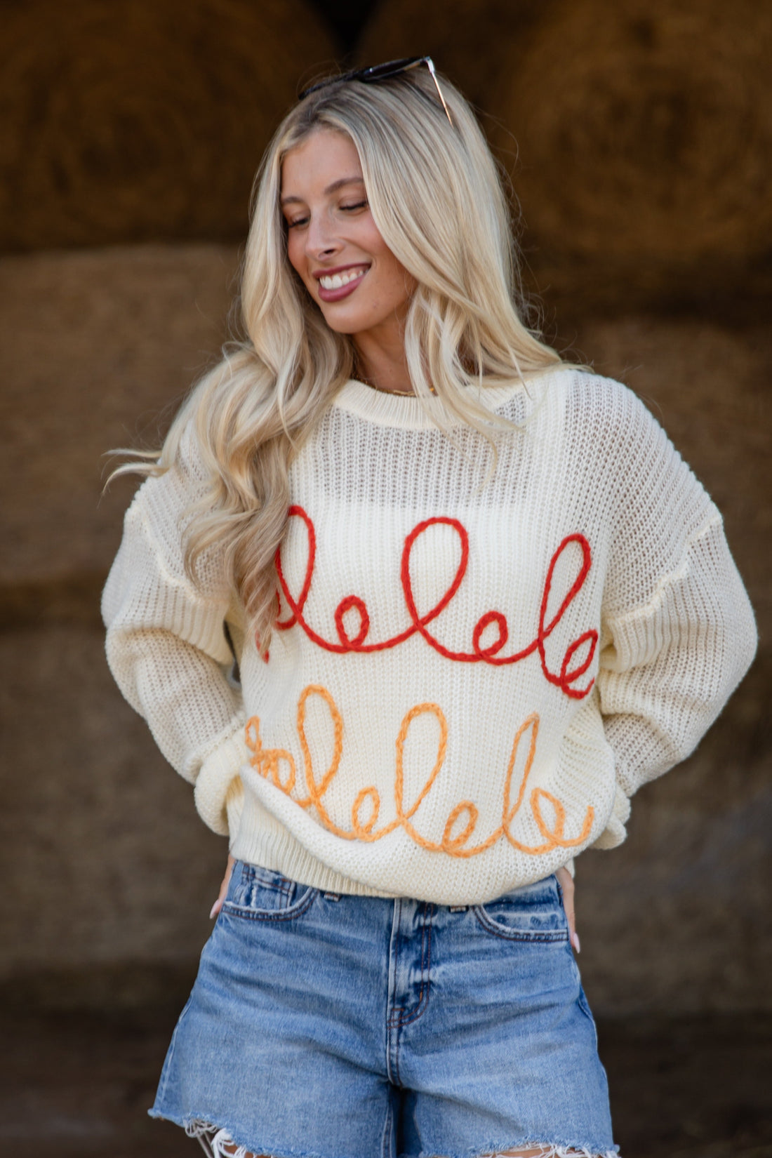 Woman wearing a cream sweater with red text and denim shorts in front of bales of hay.