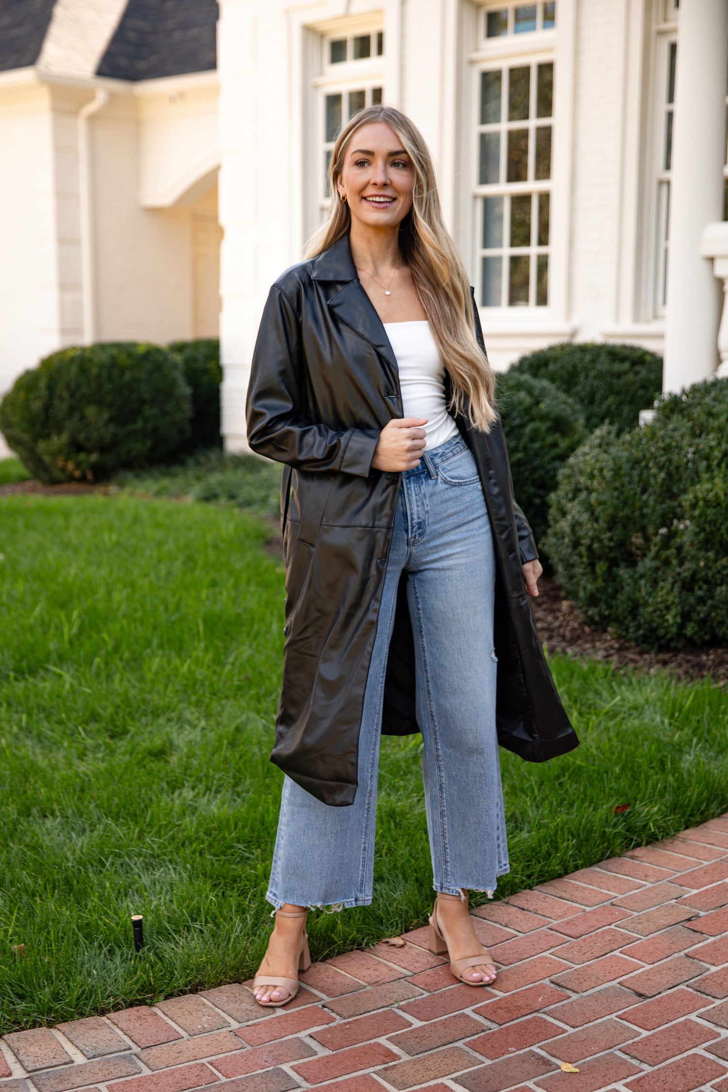 Woman standing on a brick path in front of a house wearing a black leather jacket, white top, and blue jeans.