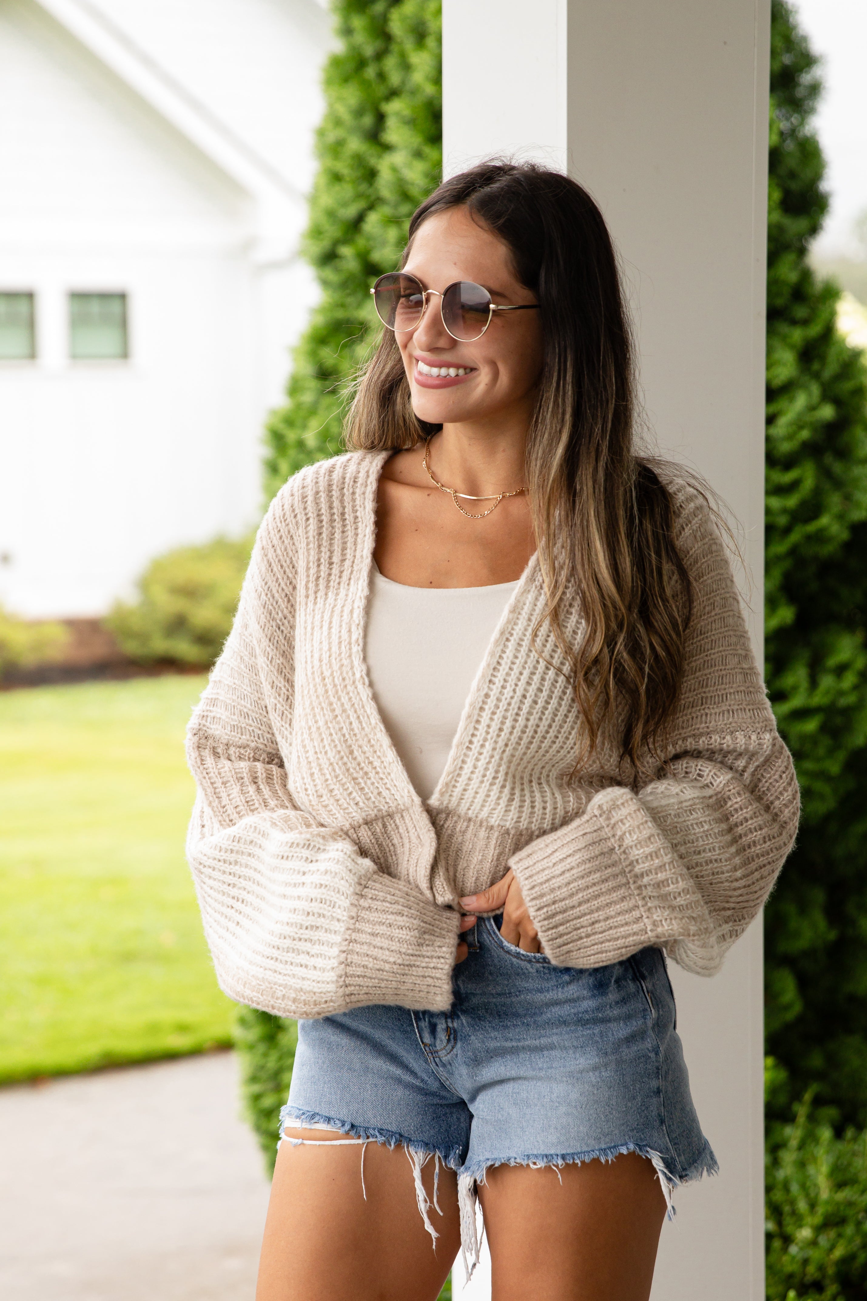 Woman wearing a beige cardigan, white top, and denim shorts outdoors.
