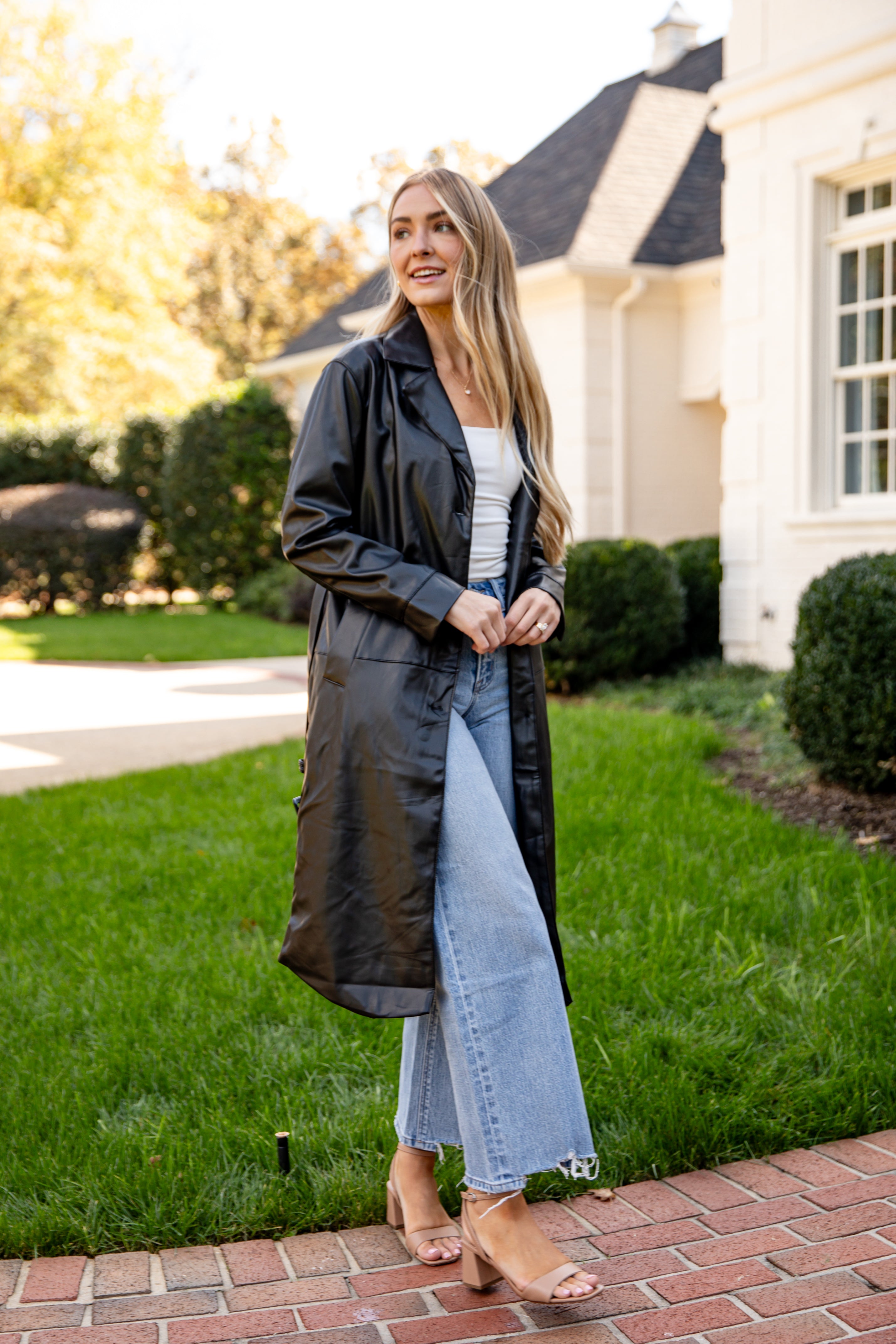 Woman standing outdoors in front of a house wearing a long black coat, white top, and light blue jeans.