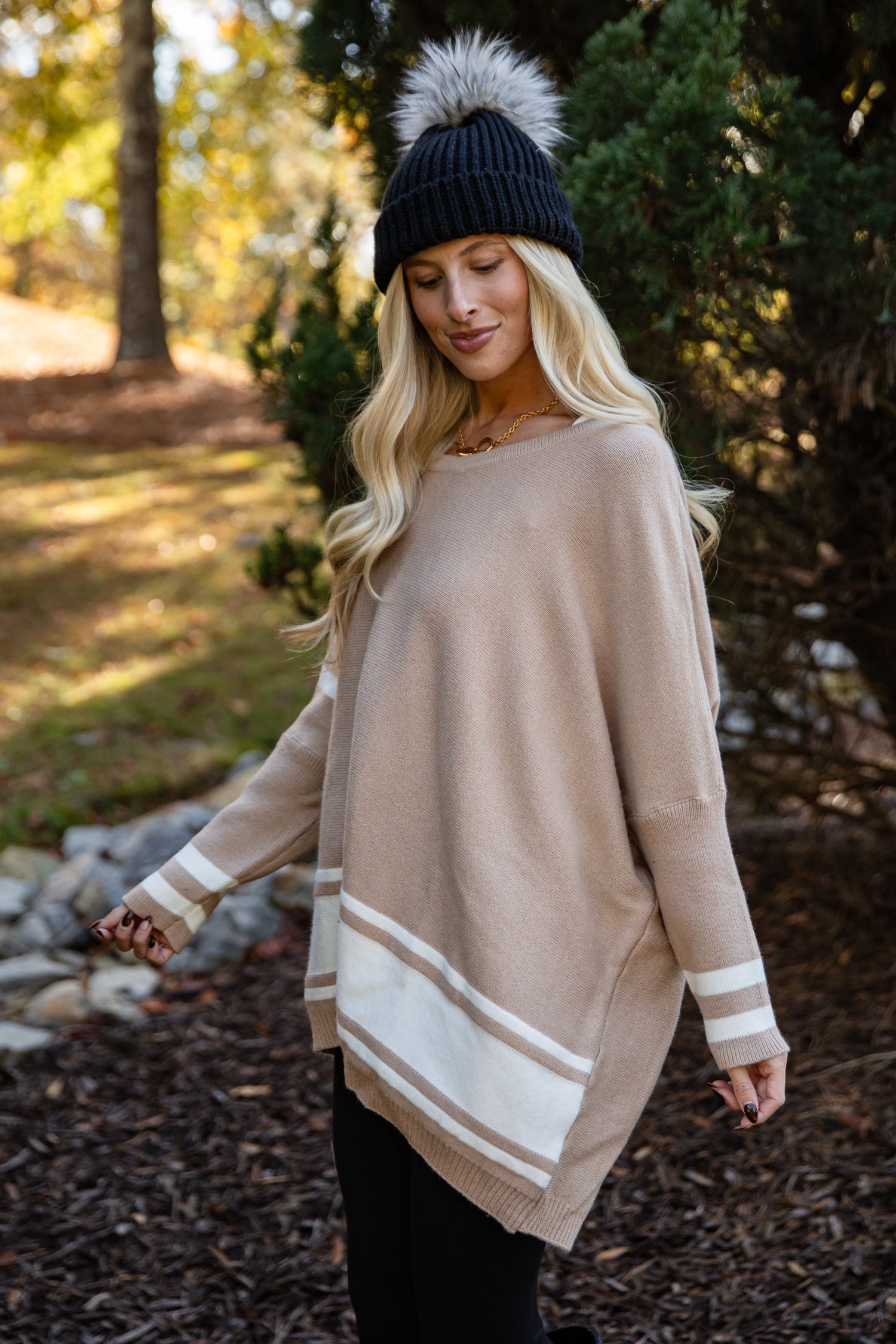 Woman wearing a beige sweater with white stripes and a black beanie in a forest setting