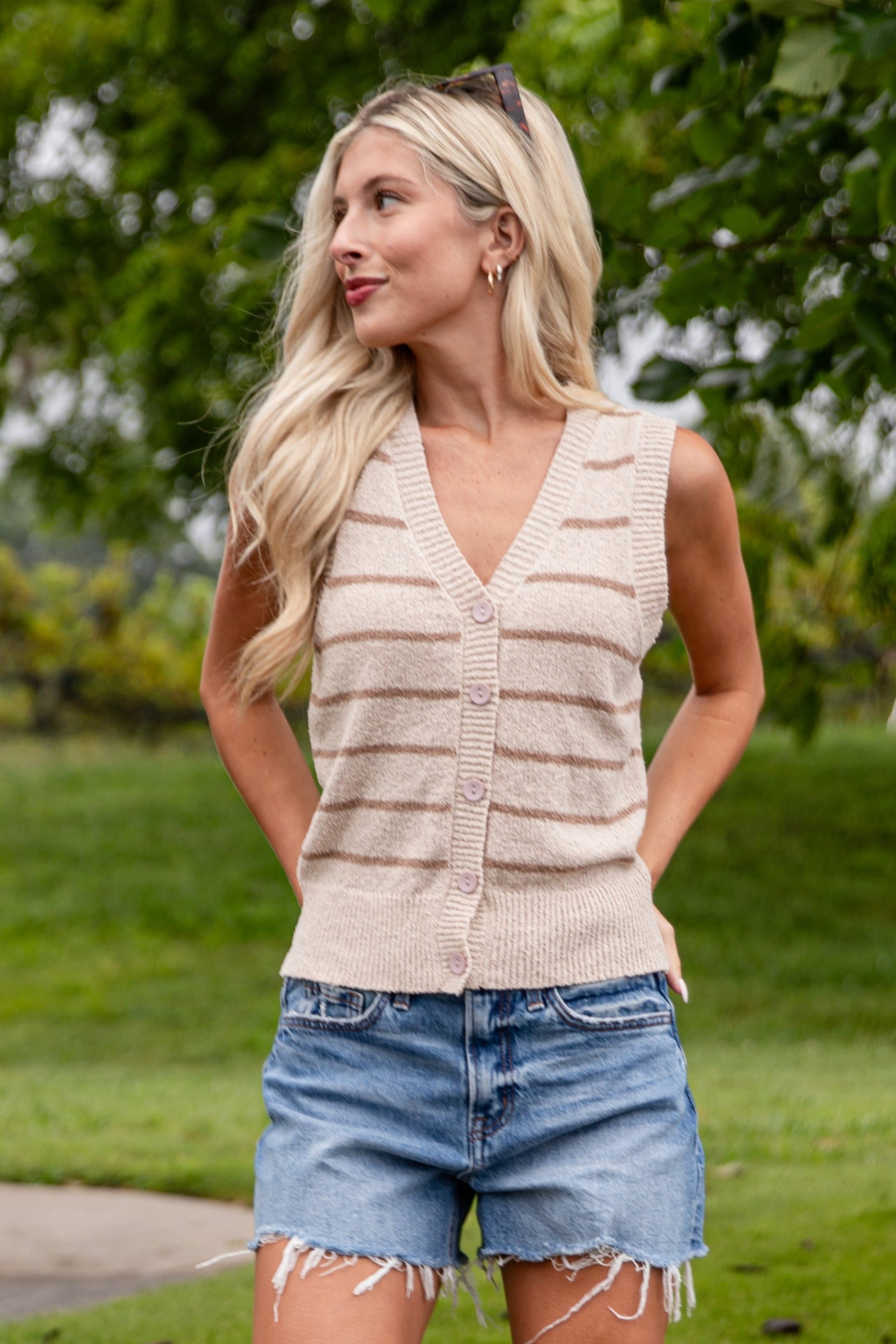 Woman wearing a beige sleeveless top, denim shorts, and beige knee-high boots standing on grass.