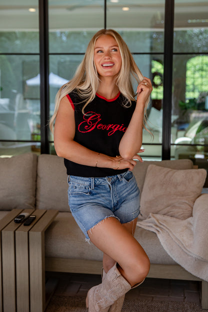 Woman sitting on a couch wearing a black sleeveless top with red text and denim shorts.