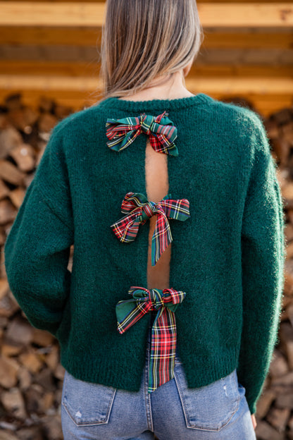 Green sweater with plaid bows on the back worn by a person, standing against a wooden background.