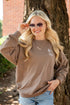A woman wearing a brown crewneck sweatshirt with a small white graphic on the left chest, paired with denim shorts, posing outdoors in front of a tree.