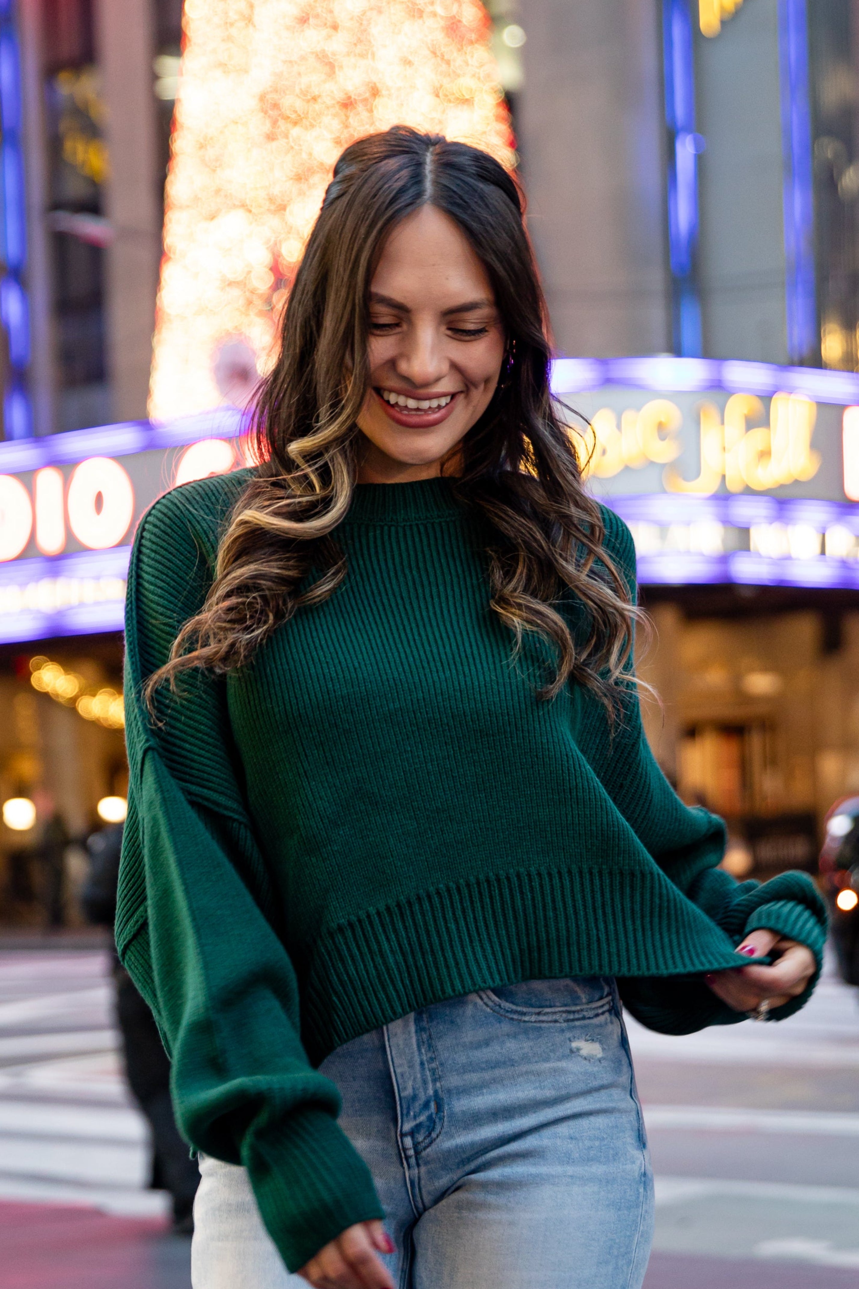 Woman in a green sweater and jeans standing on a city street with neon signs in the background