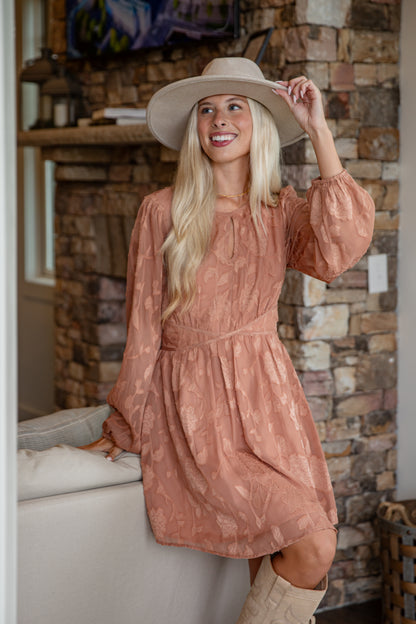Woman in a patterned dress and wide-brimmed hat standing in front of a stone wall.