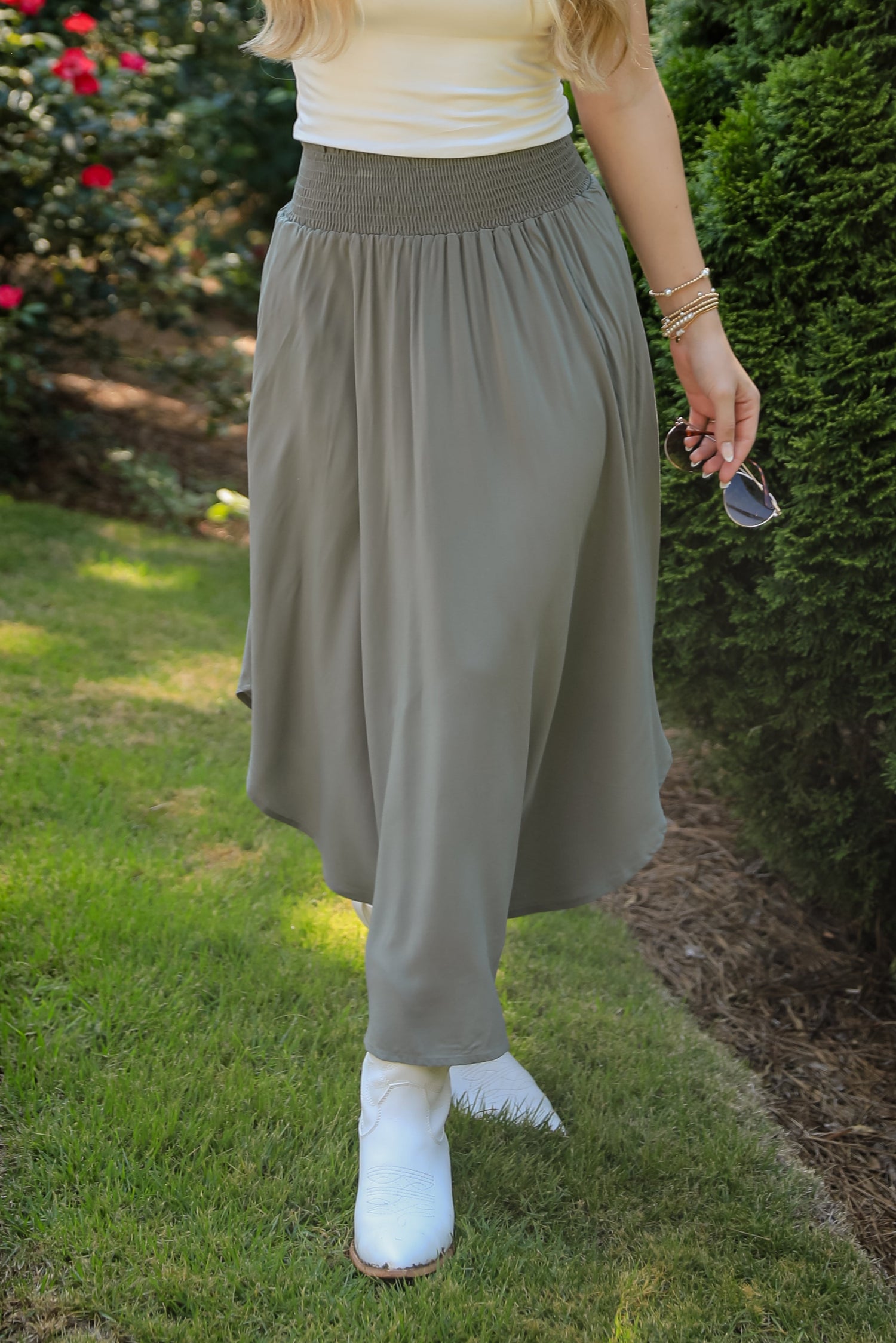 Person wearing a white top and gray skirt standing outdoors with greenery in the background
