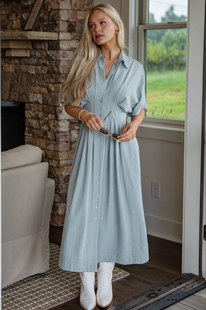 Woman in a light blue dress standing in a room with a stone fireplace and window view.