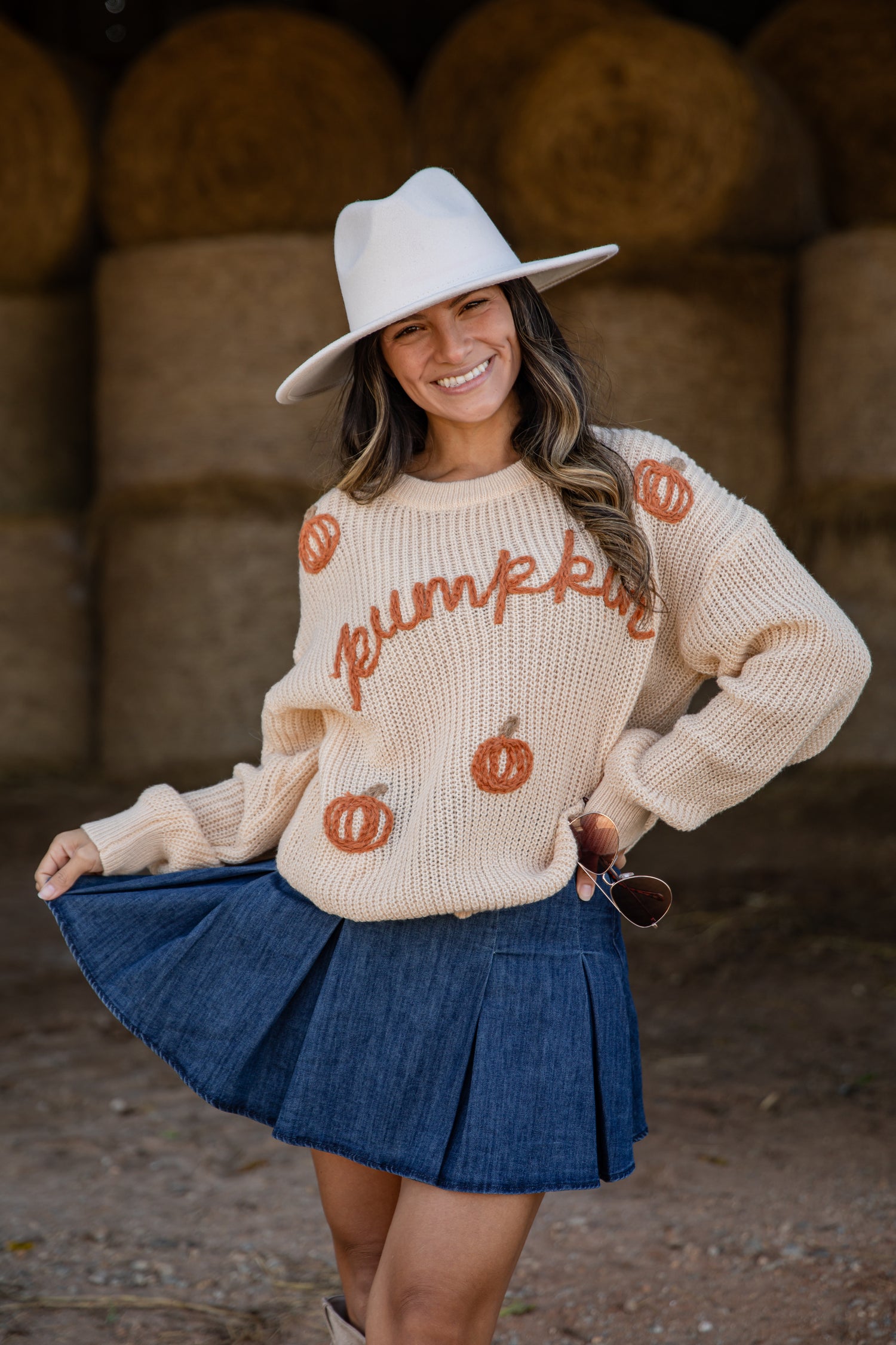 Woman wearing a cream sweater with pumpkin designs and a white hat, standing in front of hay bales.