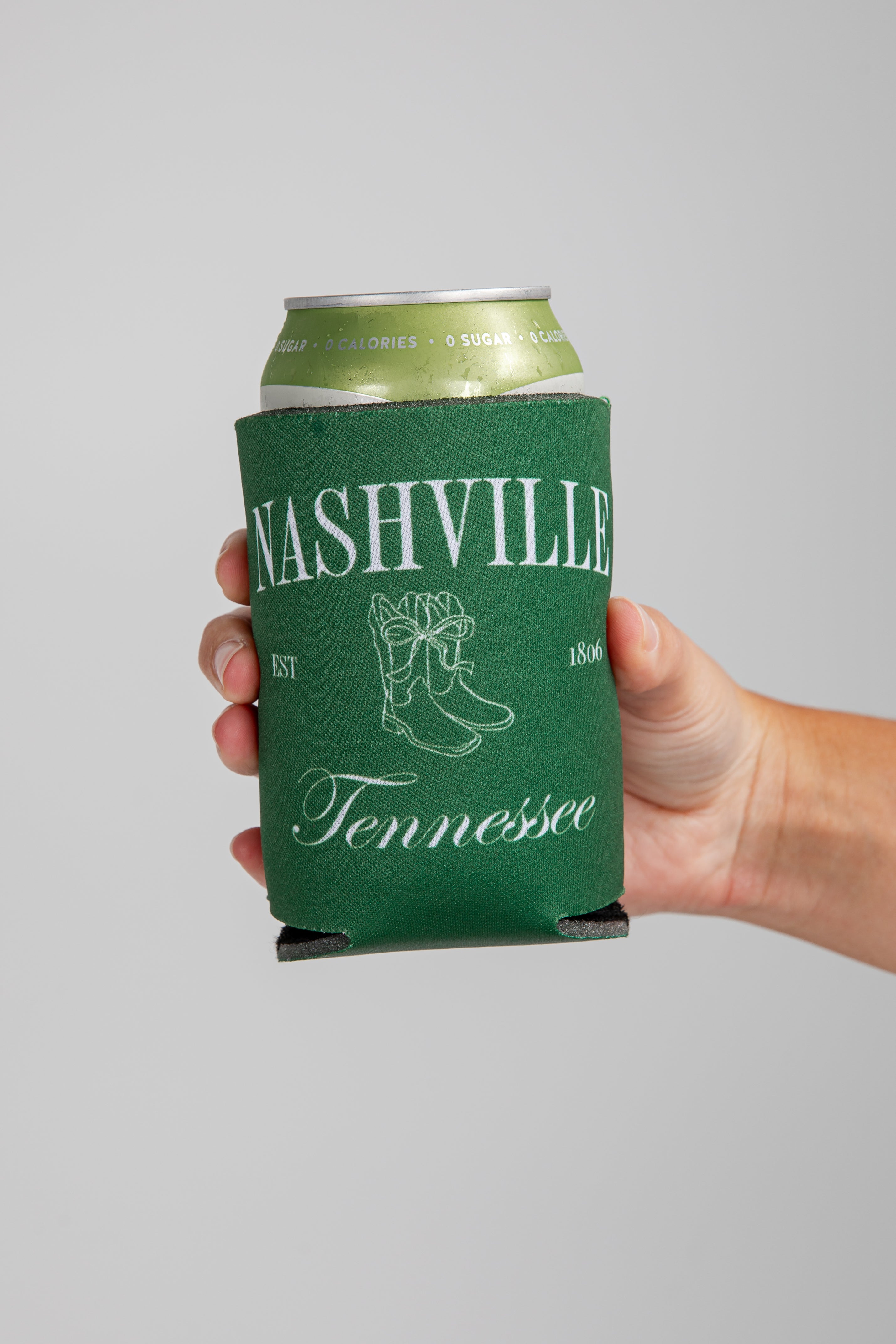 Hand holding a can with a green Nashville, Tennessee koozie against a gray background