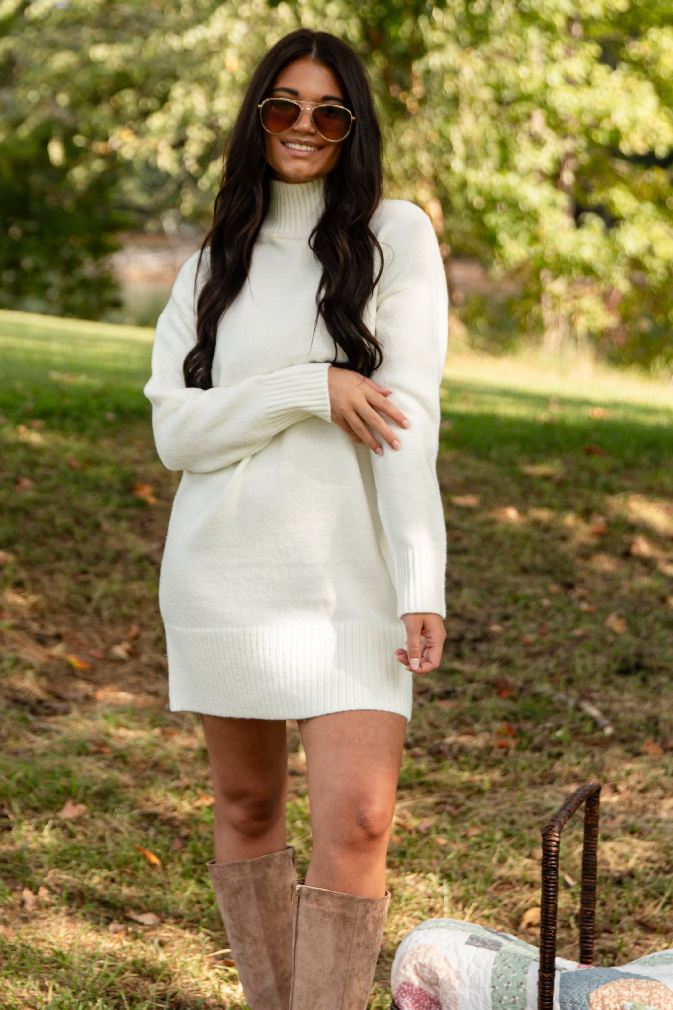 Woman in a white dress with knee-high boots standing outdoors with a picnic setup.