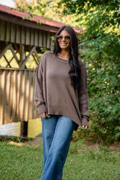 Woman wearing a brown sweater and blue jeans standing outdoors with greenery in the background