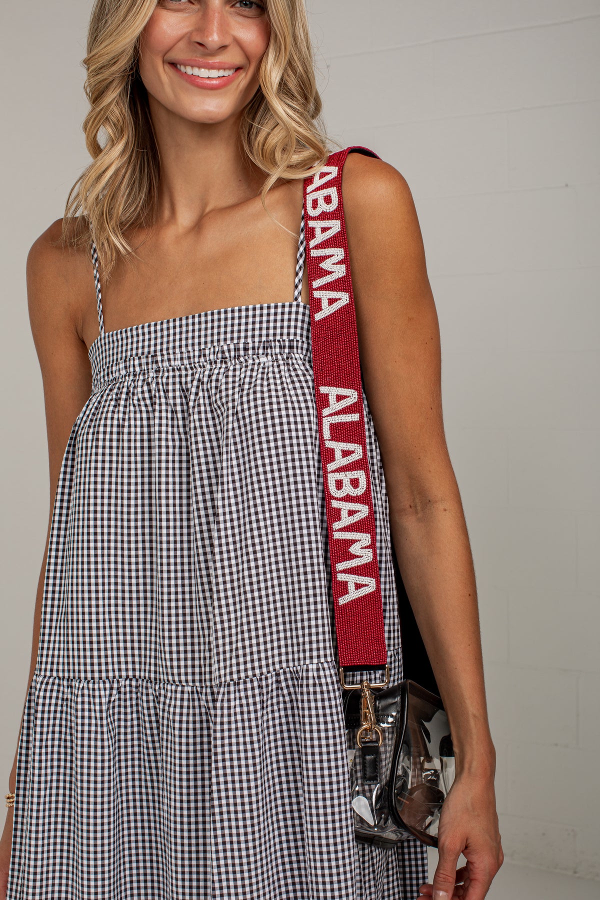 A woman wearing a black and white gingham tiered dress with a crimson red shoulder strap featuring the words &