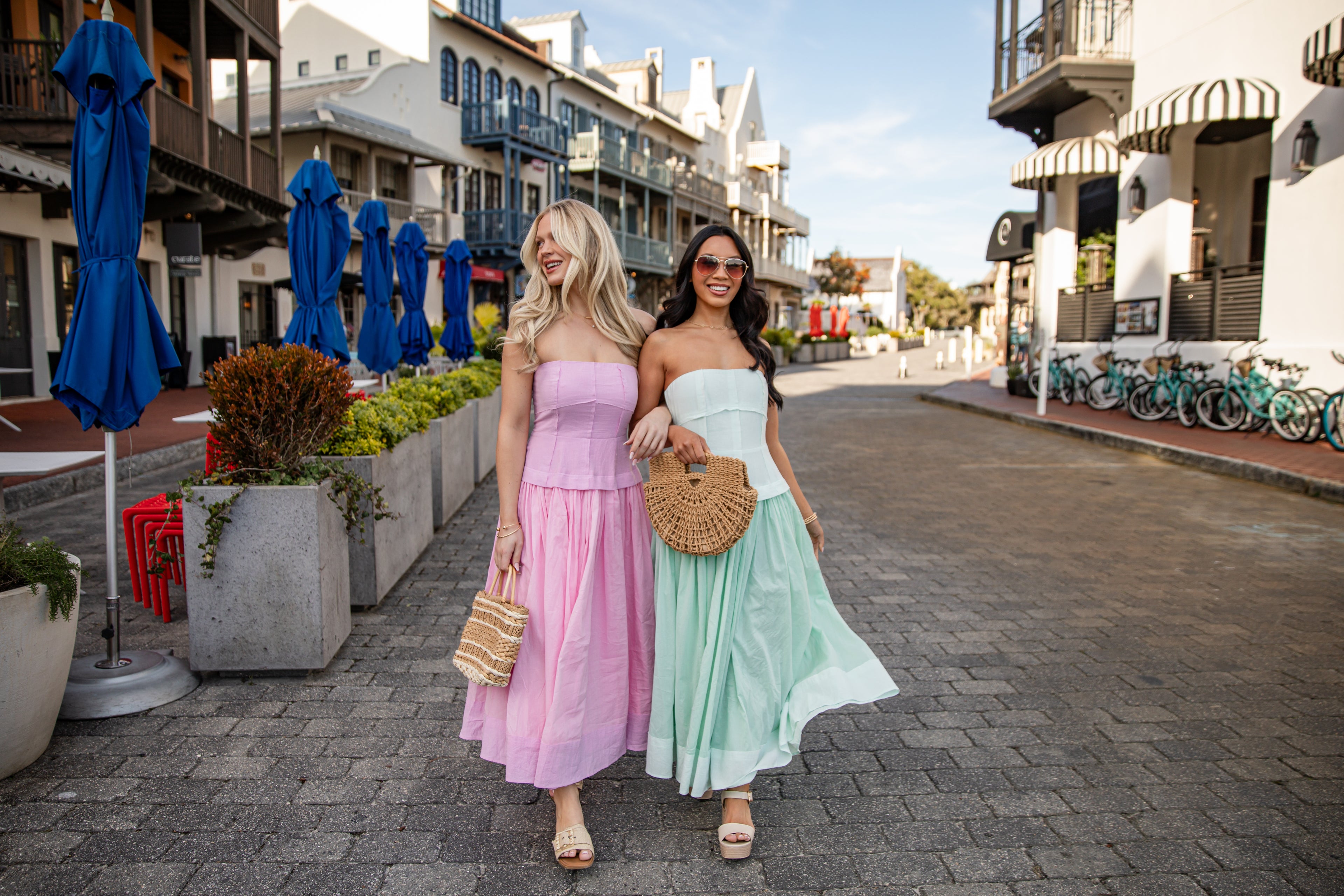 Two women in pastel dresses walking down a street with shops and bicycles in the background.