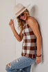 A woman wearing a brown and white buffalo check tank top, a beige wide-brim hat, and distressed blue jeans leans against a white brick wall.
