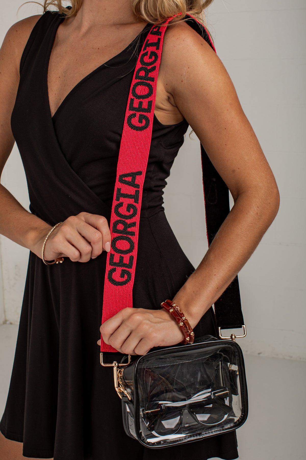 A woman wearing a black dress carries a clear plastic cosmetic bag with a bright red shoulder strap printed with the word &