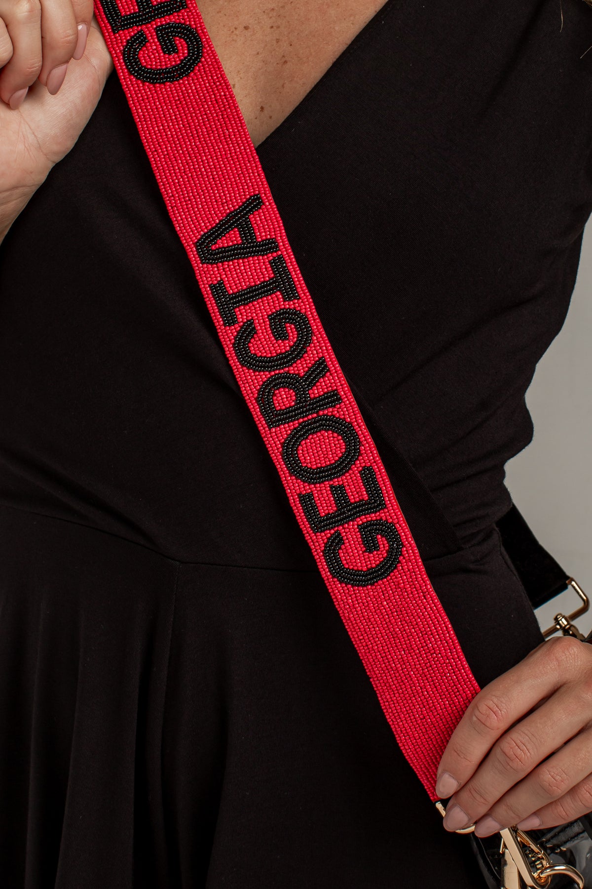 A close-up of a person wearing a black dress and holding a black handbag, with a bright red woven shoulder strap featuring the word &