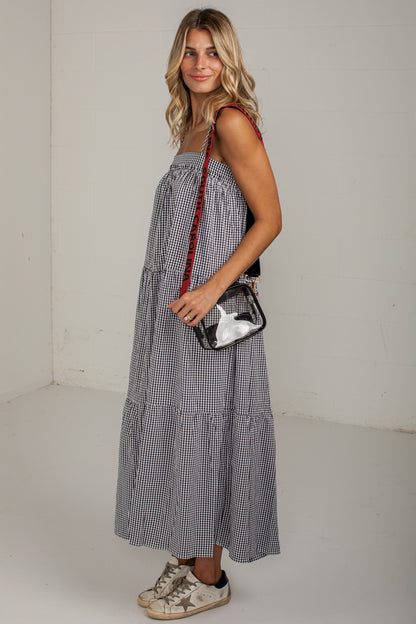 A woman modeling a sleeveless tiered maxi dress with a small black and white gingham pattern, styled with a red-and-black strap bag and distressed sneakers against a plain off-white studio background.