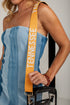 A close-up of a model wearing a light blue denim strapless top and holding a black handbag with a wide mustard yellow shoulder strap printed with the word &