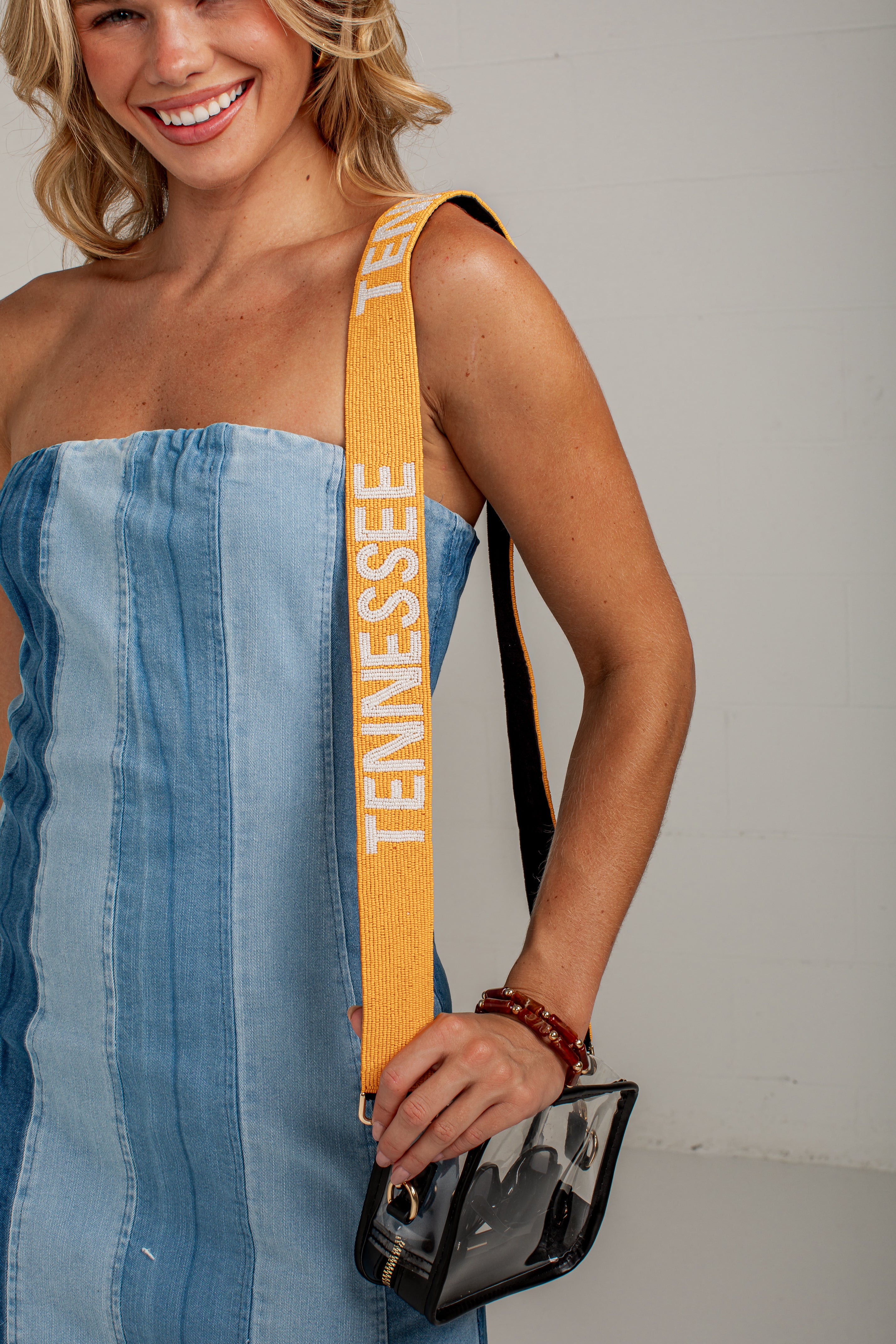 A woman wearing a blue denim strapless dress carries a small black clear handbag with a wide mustard yellow shoulder strap printed with &