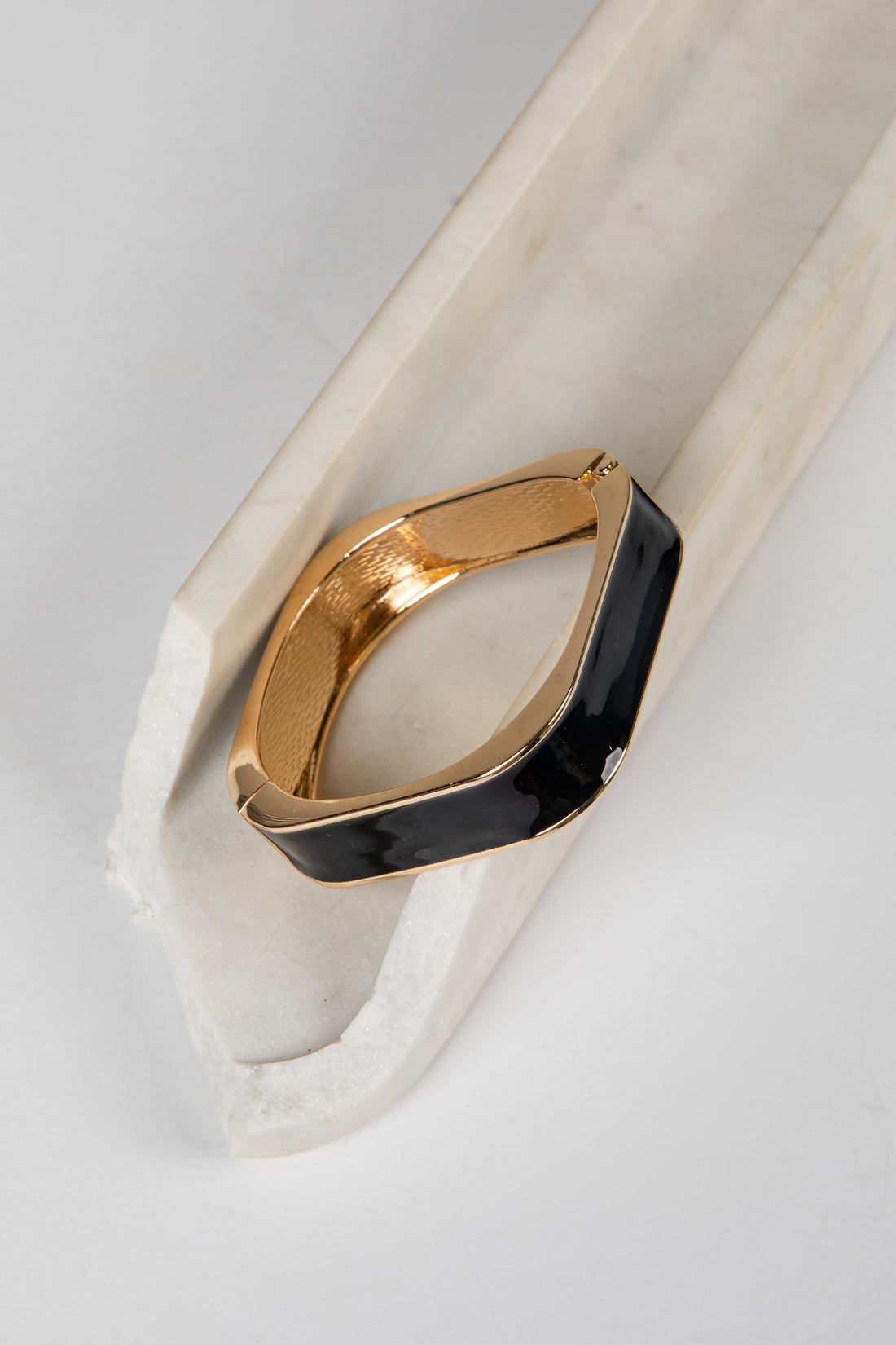 A gold-toned metal cuff bracelet with a wide, curved black enamel inlay, displayed on a white marble prop against a light gray background.