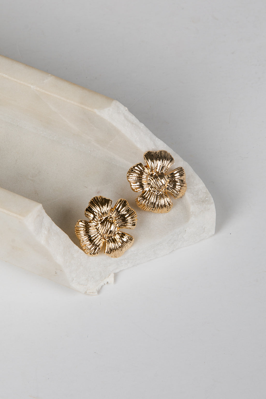 A pair of textured gold flower-shaped stud earrings resting on a white marble tray against a light gray background.