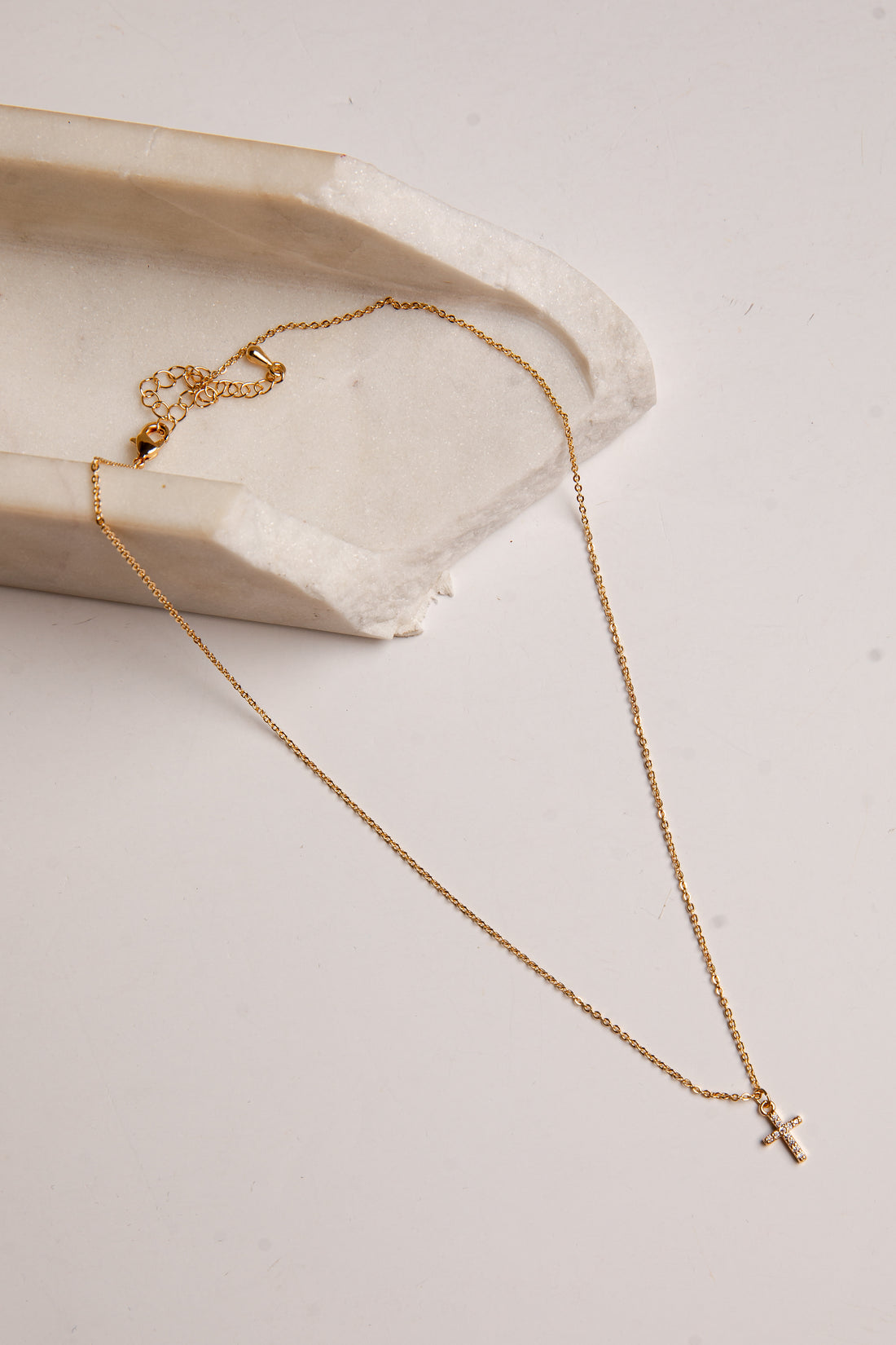 A delicate gold chain necklace with a small cross pendant, draped elegantly over a textured white stone slab on a light beige background.