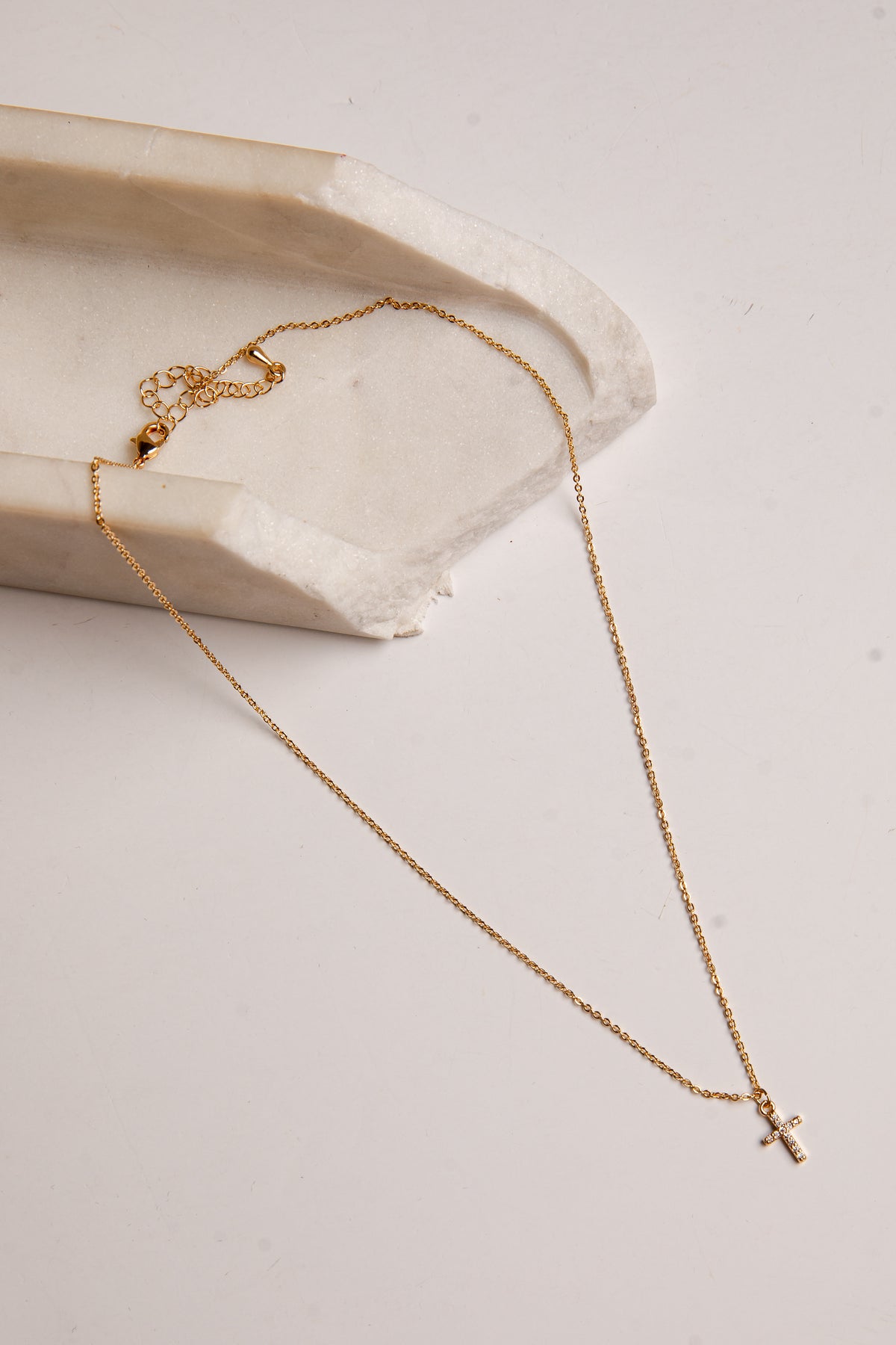 A delicate gold chain necklace with a small cross pendant, draped elegantly over a textured white stone slab on a light beige background.