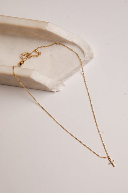 A delicate gold chain necklace with a small cross pendant, draped elegantly over a textured white stone slab on a light beige background.