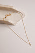 A delicate gold chain necklace with a small cross pendant, draped elegantly over a textured white stone slab on a light beige background.