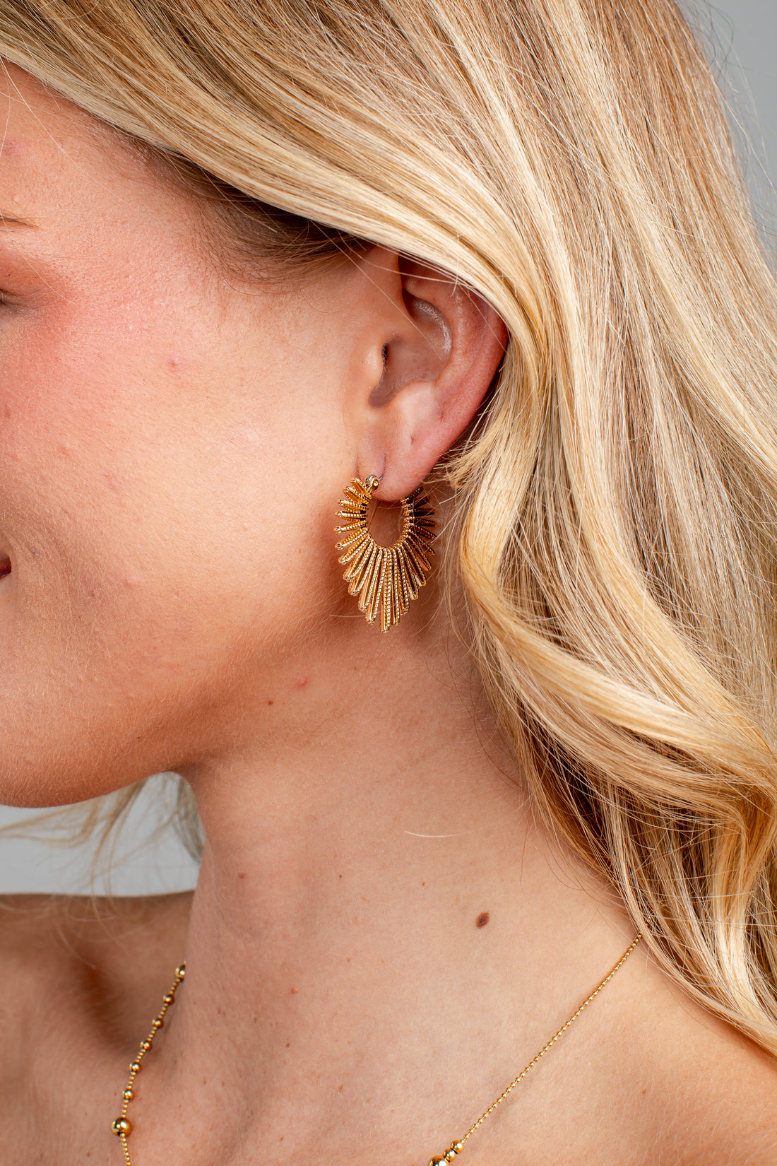 A close-up side profile of a blonde woman wearing a gold sunburst hoop earring and a delicate gold chain necklace.