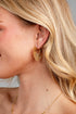 A close-up side profile of a blonde woman wearing a gold sunburst hoop earring and a delicate gold chain necklace.