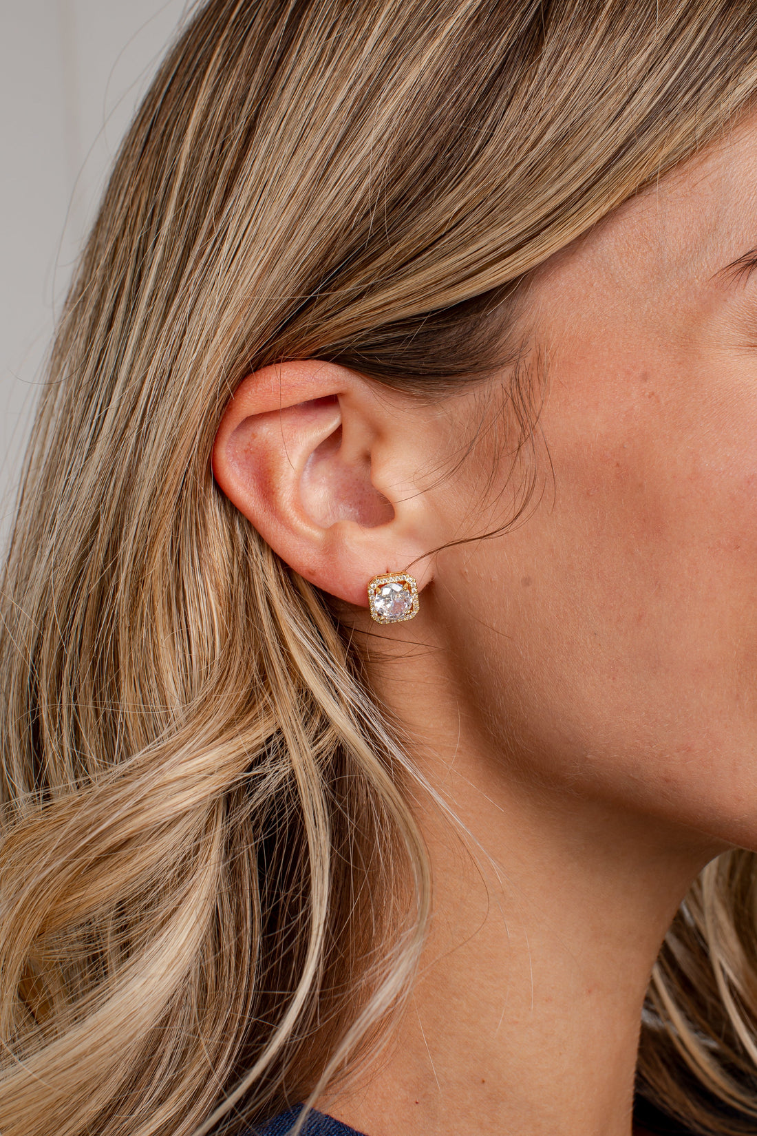 A close-up side profile of a woman with blonde hair wearing a small gold stud earring with a clear round gemstone.