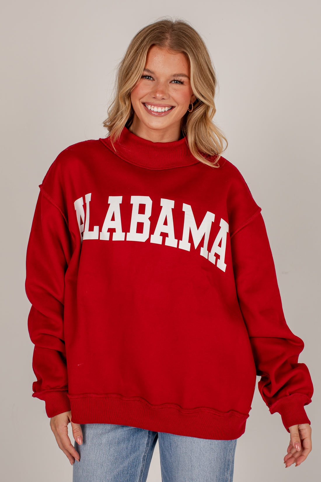 A smiling blonde woman wearing a red mock-neck sweatshirt with &