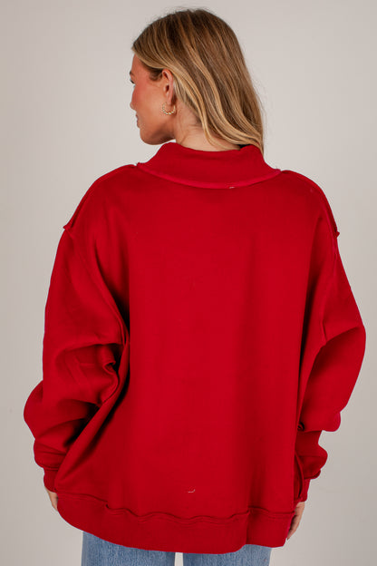 A woman viewed from the back wearing an oversized bright red sweatshirt with a mock neck and distressed sleeve edges, paired with blue jeans against a plain light gray studio background.